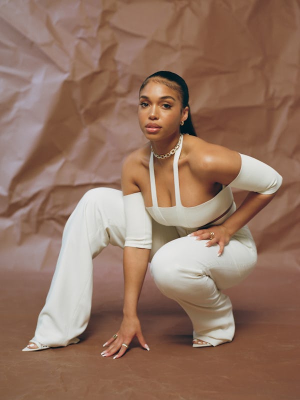 Model Lori Harvey poses against a brown backdrop with one hand on the ground and wears a white cut-o...