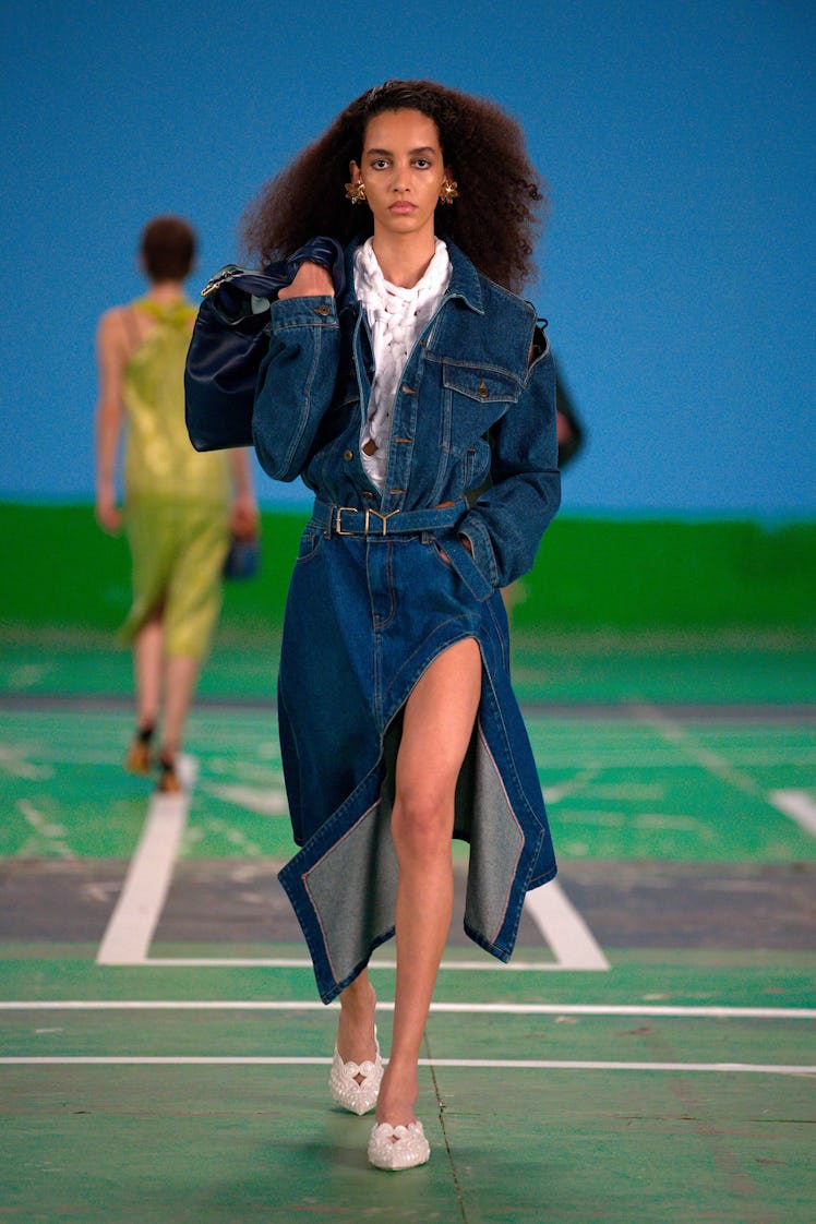 A curly-haired female model walking while wearing a denim dress over a white shirt