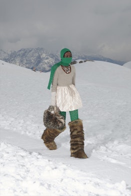 Model walking in the snow carrying a furry bag and wearing big furry boots and a green knitted scarf...