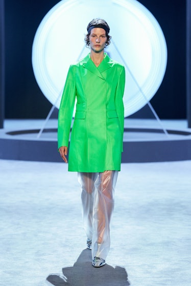 Model walking down the runway wearing see-through pants and a long green leather coat