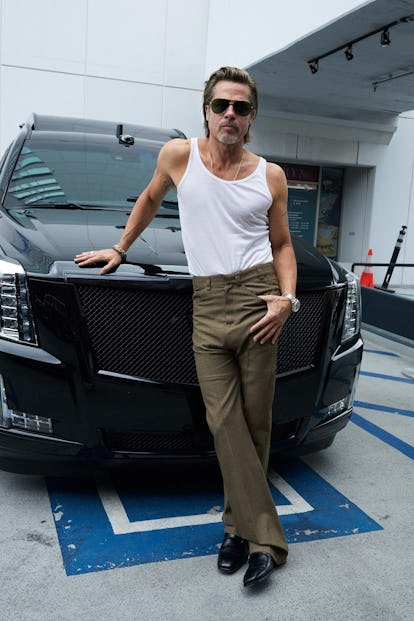 Brad Pitt standing in front of a black SUV wearing sunglasses, a white tank top, and sage green pant...