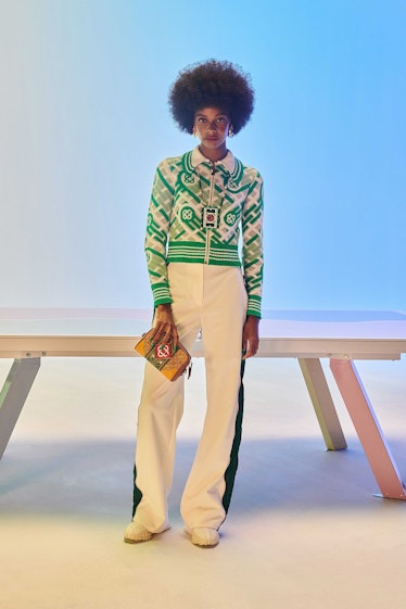 A curly-haired female model posing while wearing a green sweater and white pants
