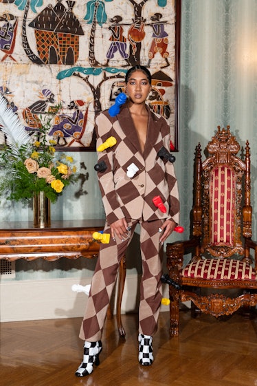 A  model in a beige and brown checkered Pyer Moss suit with large multicolored chess pieces as acces...