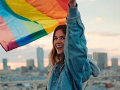 Young woman waving Pride flag, wearing a jean jacket in need of a rainbow quote, caption, or pun for...