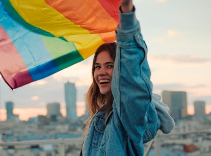 Young woman waving Pride flag, wearing a jean jacket in need of a rainbow quote, caption, or pun for...