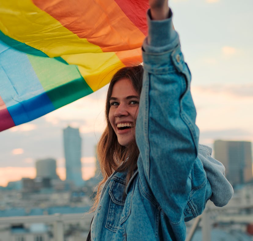 Young woman waving Pride flag, wearing a jean jacket in need of a rainbow quote, caption, or pun for...