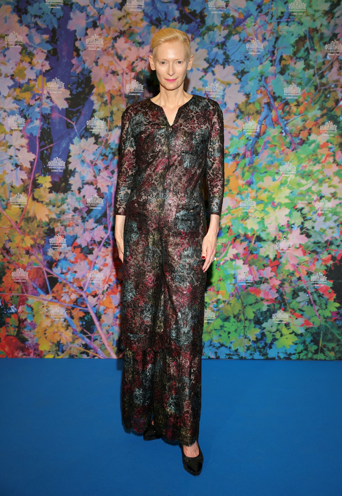 Tilda Swinton wearing shiny suit against multicolor floral background