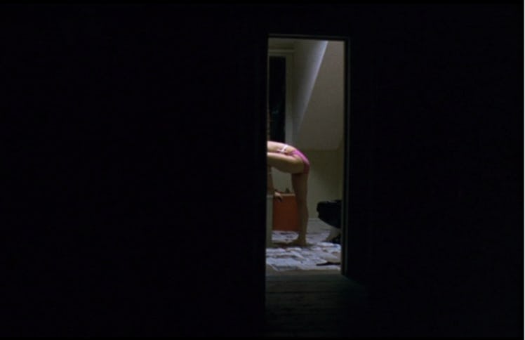 Zoe Lister-Jones visible through a door in a dark room with one leg raised bending over