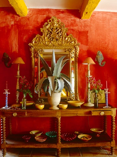 A room with red walls, a golden-framed mirror and plants and bowls on a wooden table