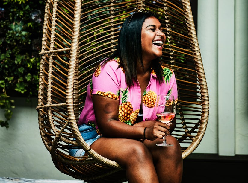 Young woman laughing in a hanging chair, holding a glass of wine before posting a pic on Instagram w...