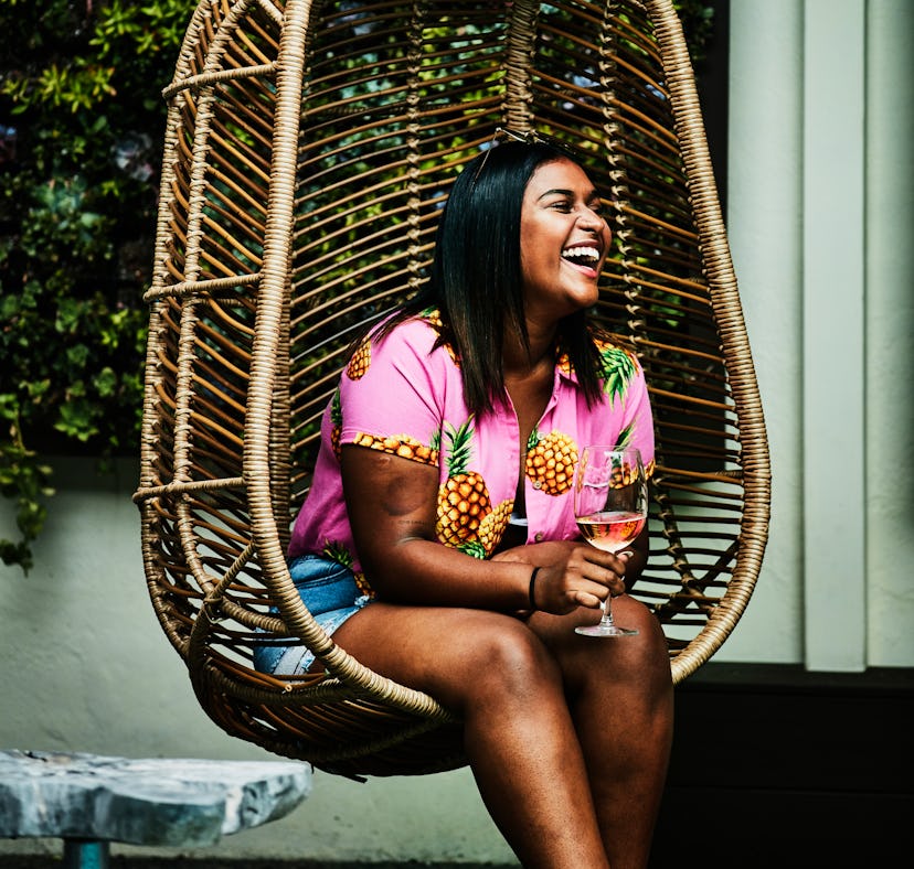 Young woman laughing in a hanging chair, holding a glass of wine before posting a pic on Instagram w...