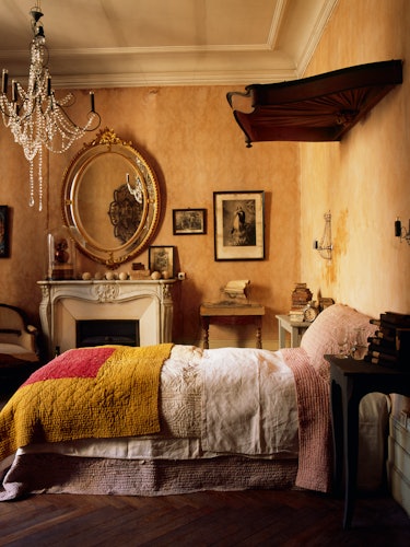 A bedroom with yellow walls, a fireplace, an oval mirror and a bed with layered bedsheets