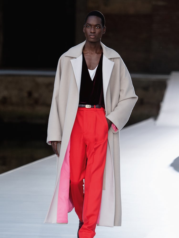 A model in a beige coat, black top and red pants at the Valentino Couture Fall 2021 Couture