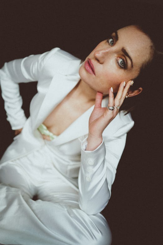 A portrait of Zoe Lister-Jones posing in a white suit and a white hand-shaped top