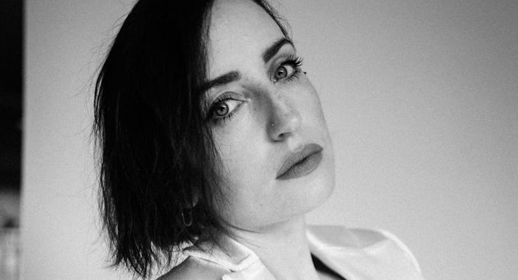 A portrait of Zoe Lister-Jones posing in a white blazer and a white hand-shaped top