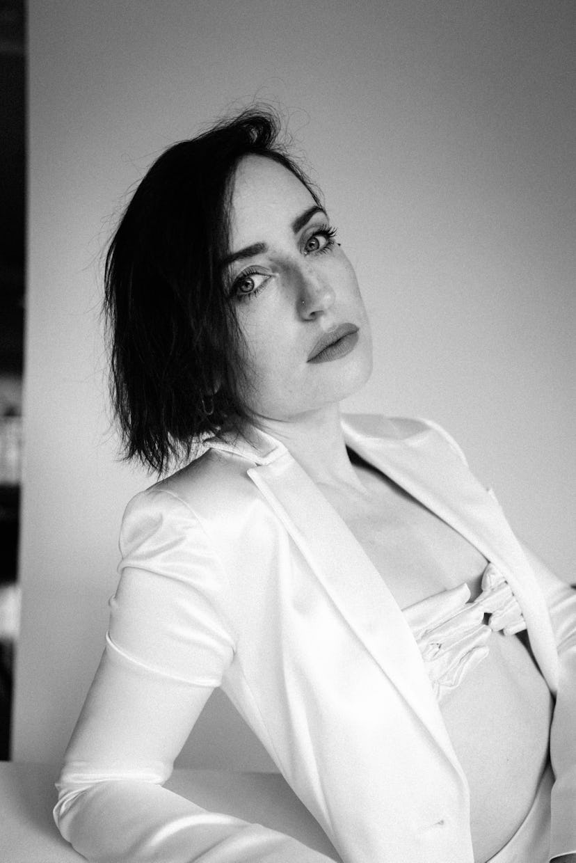 A portrait of Zoe Lister-Jones posing in a white blazer and a white hand-shaped top