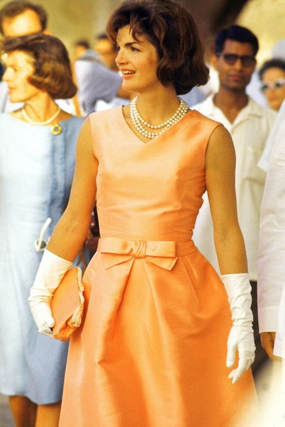 Jackie Kennedy wearing a peach dress with a bow at the waist, holding a peach bag with white gloves ...
