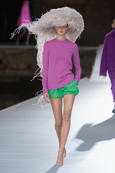 A model walking the runway in a purple sweater and green shorts with a white head piece by Valentino...
