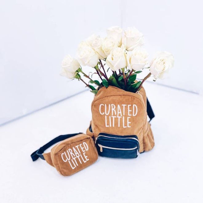 Brown fanny pack and brown bookbag that say "curated life"; bookbag is holding white flowers 