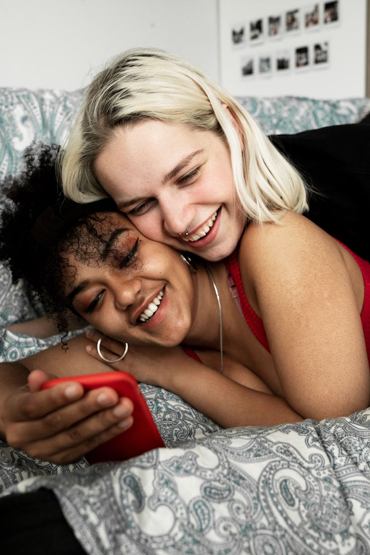 2 smiling young women laying in bed, looking at a phone deciding what one-year anniversary caption t...