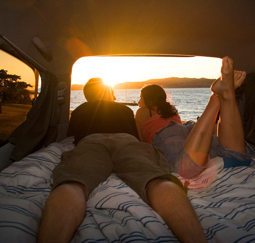 Couple watching sunset from bed inside campervan before posting on Instagram with romantic sunset qu...
