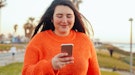 Young woman wearing an orange oversized sweater, looking at her phone trying to figure out which swe...