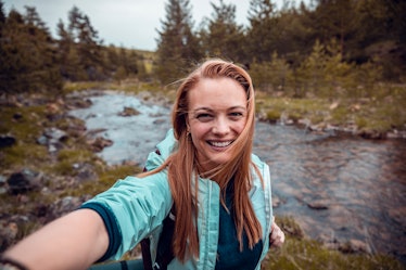 Young woman taking a selfie in front of a stream on a hike before posting the pic with camping Insta...
