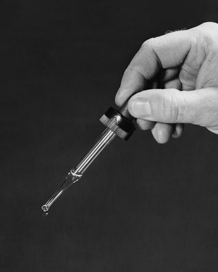 A black and white image of a hand holding a pipette with a drop of Facial Oil dripping.