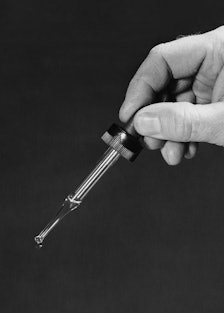 A black and white image of a hand holding a pipette with a drop of Facial Oil dripping.