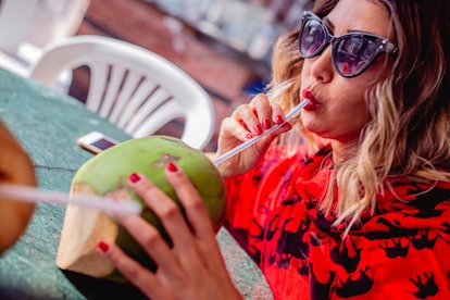 Young woman sipping out of a coconut before posting on Instagram with coconut puns, coconut quotes, ...