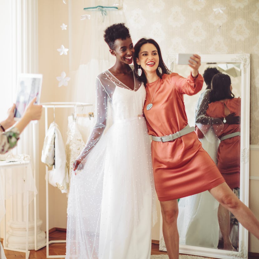 Young bride trying out her wedding dress, taking a selfie with her friend before posting on Instagra...