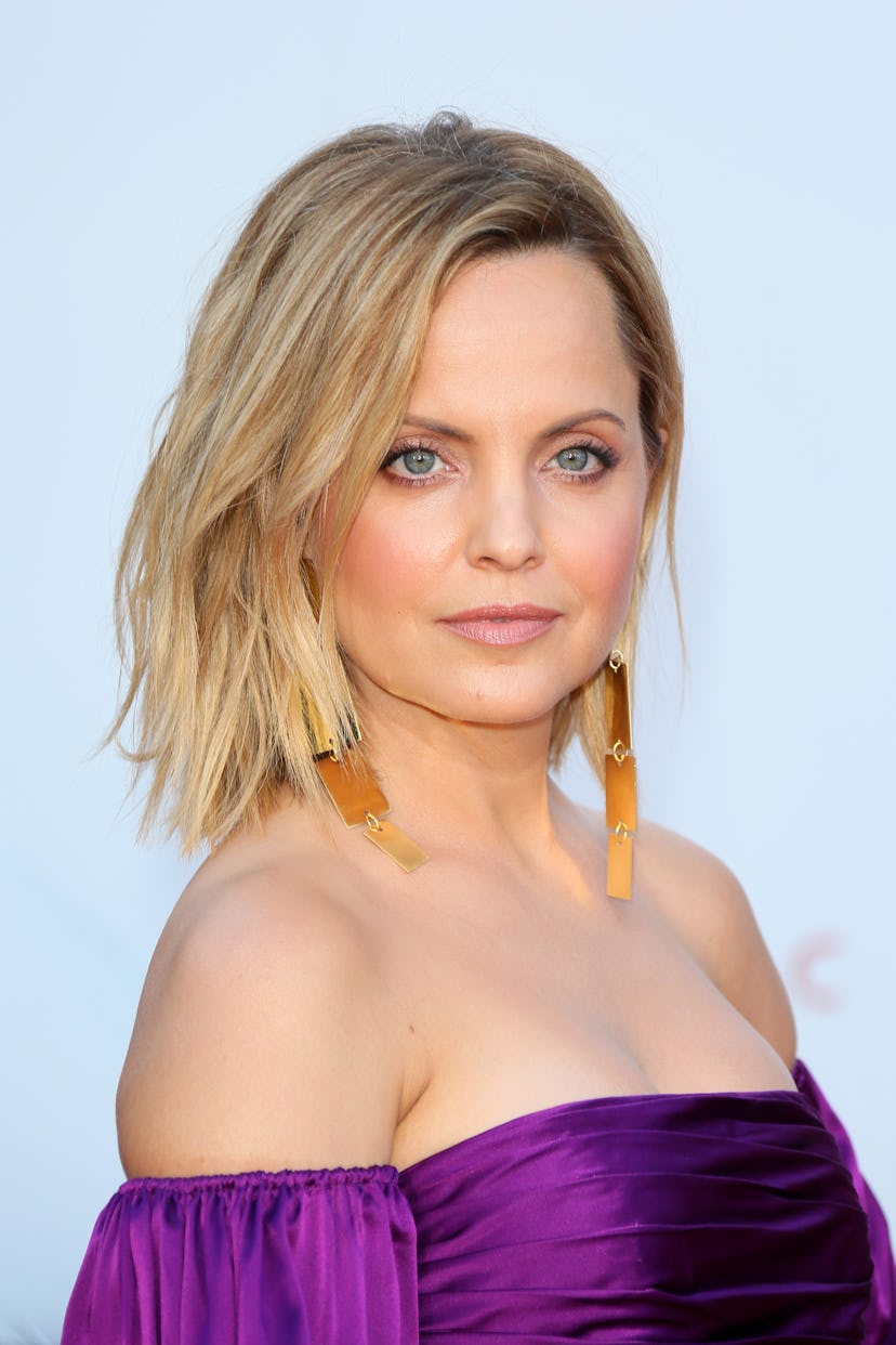A portrait of Mena Suvari in a purple off-the-shoulder dress and gold earrings