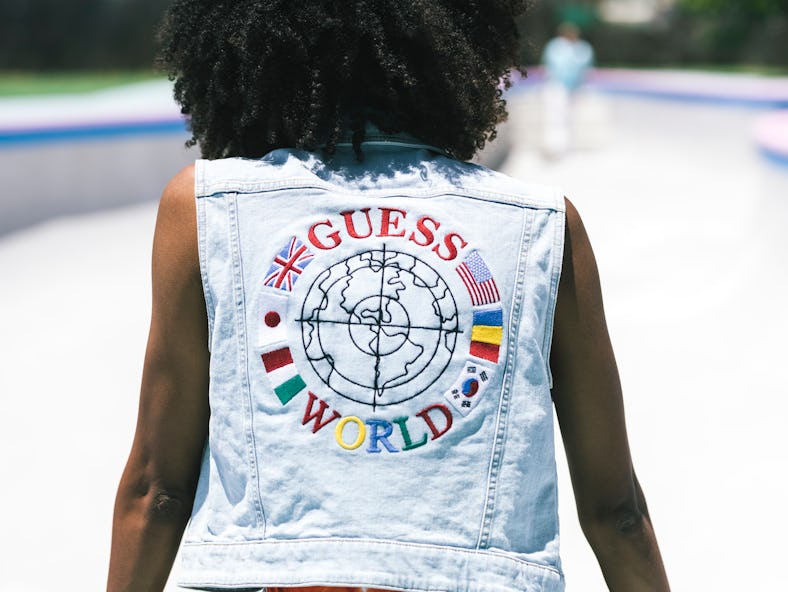 A Black woman with natural hair wearing a light wash denim jacket with a Guess World graphic from it...