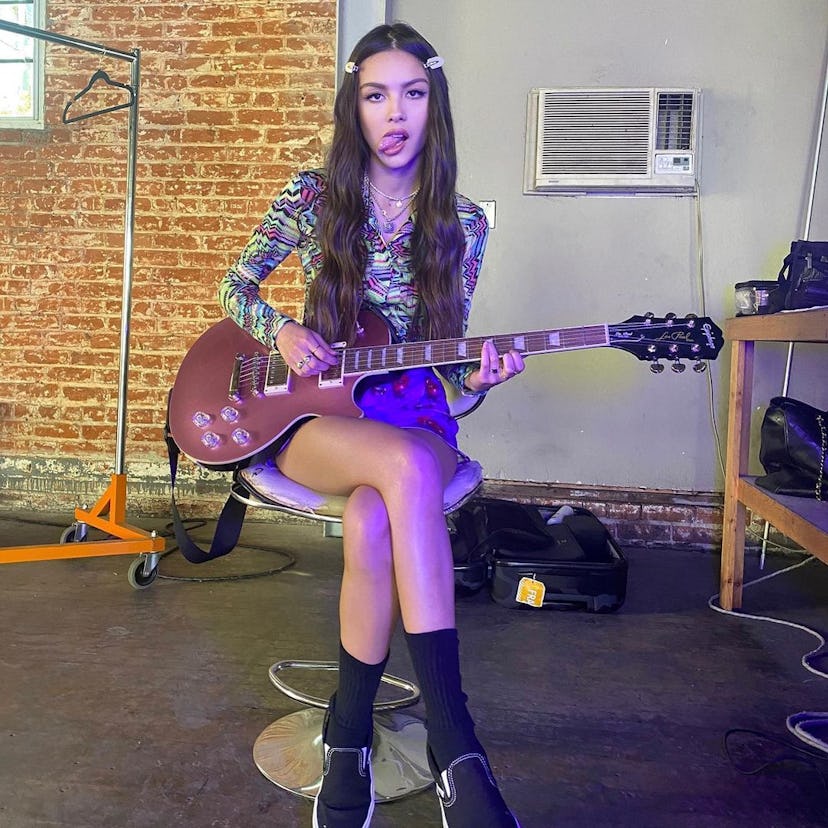 Olivia Rodrigo sitting on a stool holding a guitar and wearing a long-sleeve blouse, printed mini sk...