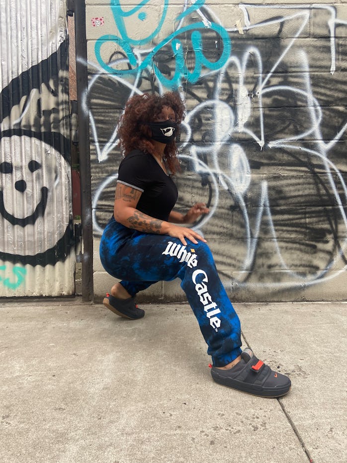 The author poses in front of a graffitied wall wearing blue and black sweatpants that say White Cast...