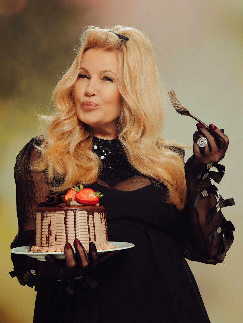 Jennifer Coolidge in a black Gucci dress and gloves, holding a chocolate strawberry cake and fork 