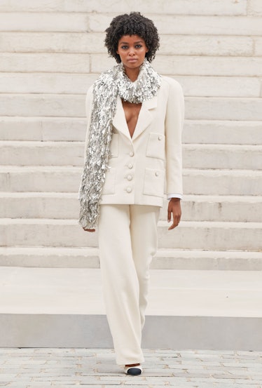 A model walking in a white Chanel suit and silver shimmery scarf with white and black flats 