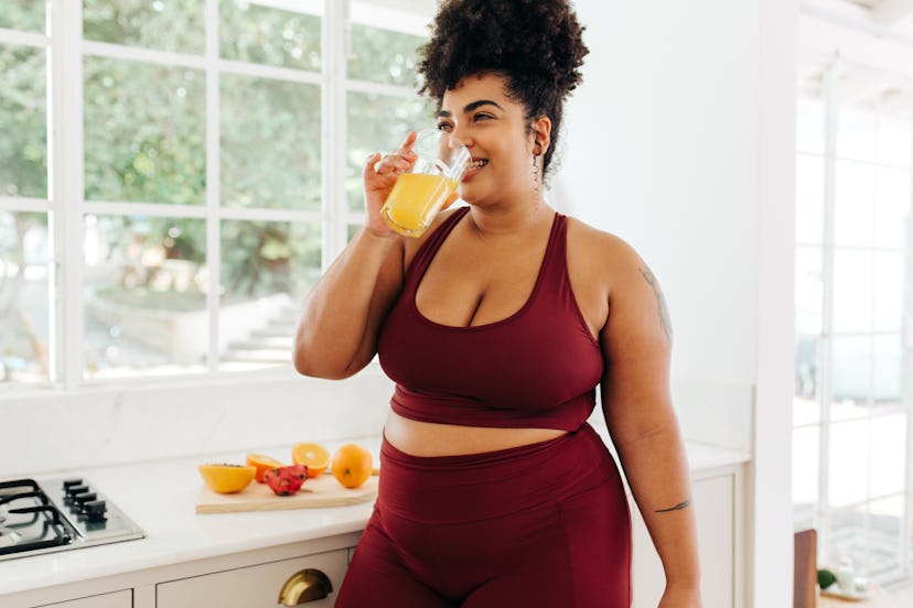A woman wearing a burgundy yoga set drinking pineapple juice while standing in a kitchen