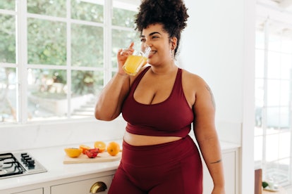 A woman wearing a burgundy yoga set drinking pineapple juice while standing in a kitchen