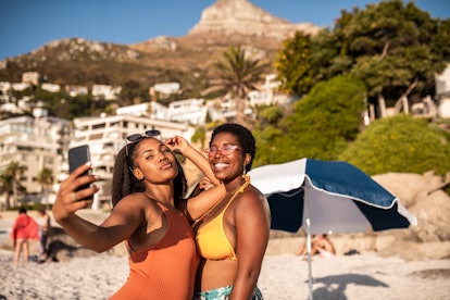 2 friends taking a selfie at the beach in the summer sun before posting a pic on Instagram with a fu...