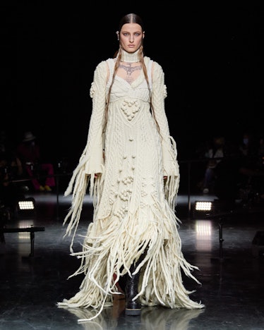 A model on the runway in a white, crochet-looking Jean Paul Gaultier gown with long sleeves and frin...