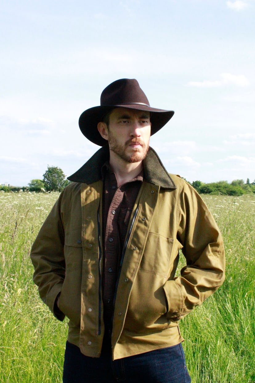Andrew Koji standing in a field in a brown shirt, a khaki jacket and a brown hat
