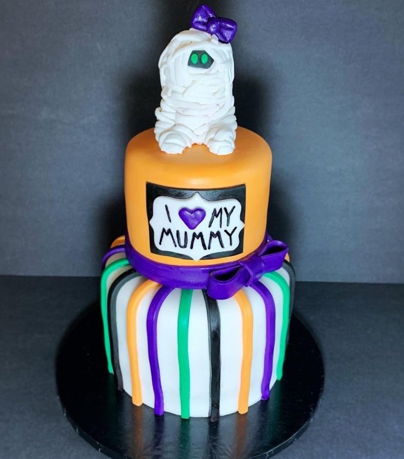 Two tiered cake with baby mummy topper; decorated in Halloween colors orange, black, green, and purp...