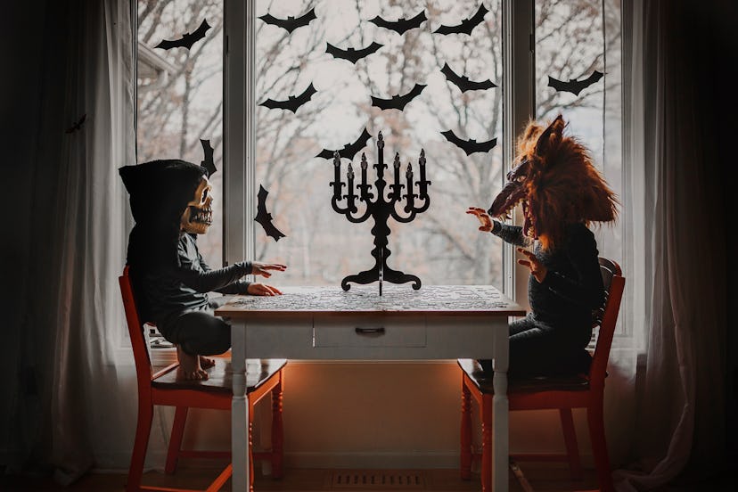 Two kids wearing Halloween masks, sitting at a table with a candelabra, in front of a window with ba...