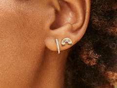 A black model with short, natural hair texture models two dainty pieces of jewelry: a rhinestone hoo...