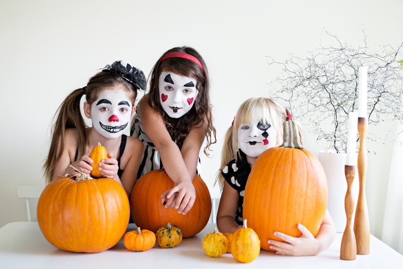 Three girls sitting at table with pumpkins, their faces painted like clowns in black, white, and red...