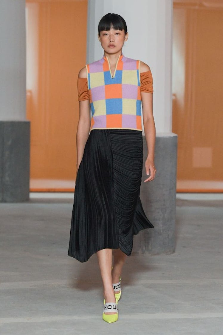 A model walking the runway in a black skirt and checkered vest by Stine Goya