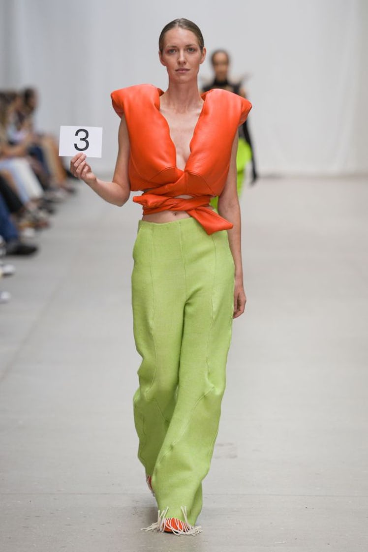 A model walking in look from Emilie Axters’s collection with an orange puffy top and green pants 