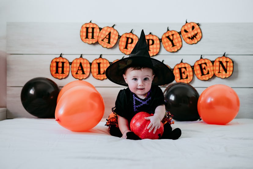Baby dressed up in witch costume, sitting on bed with black and orange balloons on either side and "...