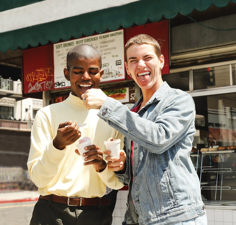 Two young males being playful while eating ice cream in front of a cafe during the day, waiting to p...